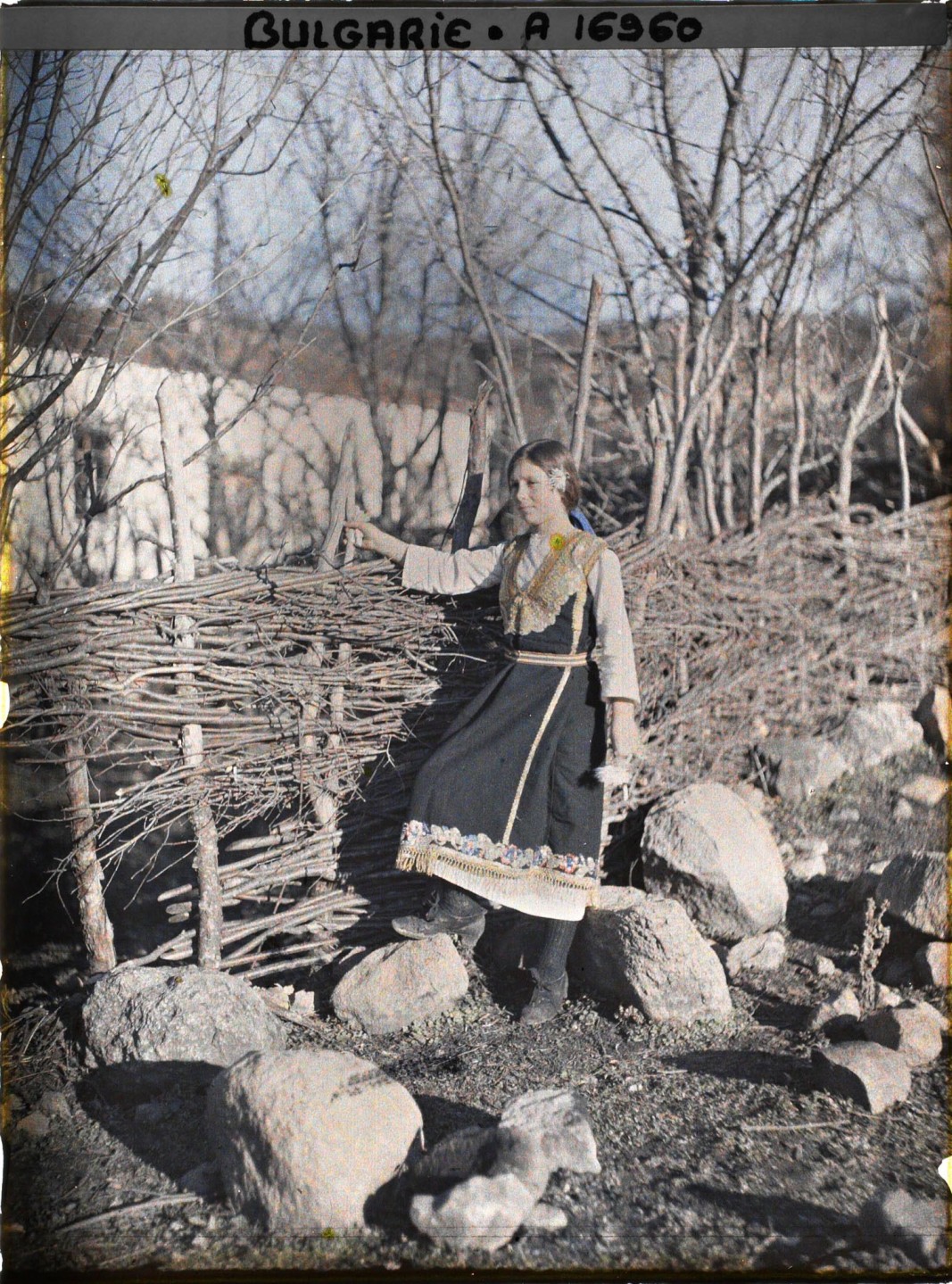 Село Владая в покрайнините на София, България, Младо момиче в бродирана носия пред плет, на заден план се вижда църква