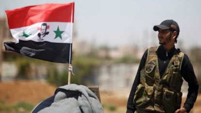 Сирийски войник до развято национално знаме в провинция Дераа, която преминава под контрола на силите на режима в Дамаск.