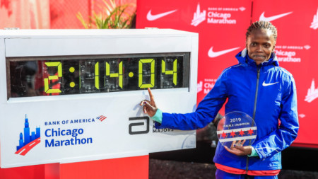 Косгеи позира пред таблото със световния си рекорд в маратона, постигнат в Чикаго.