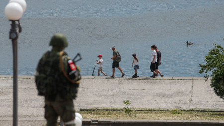 Руски военнослужещ стои на стража, докато семейство се разхожда край р. Днепър в Херсон, май 2022 г.